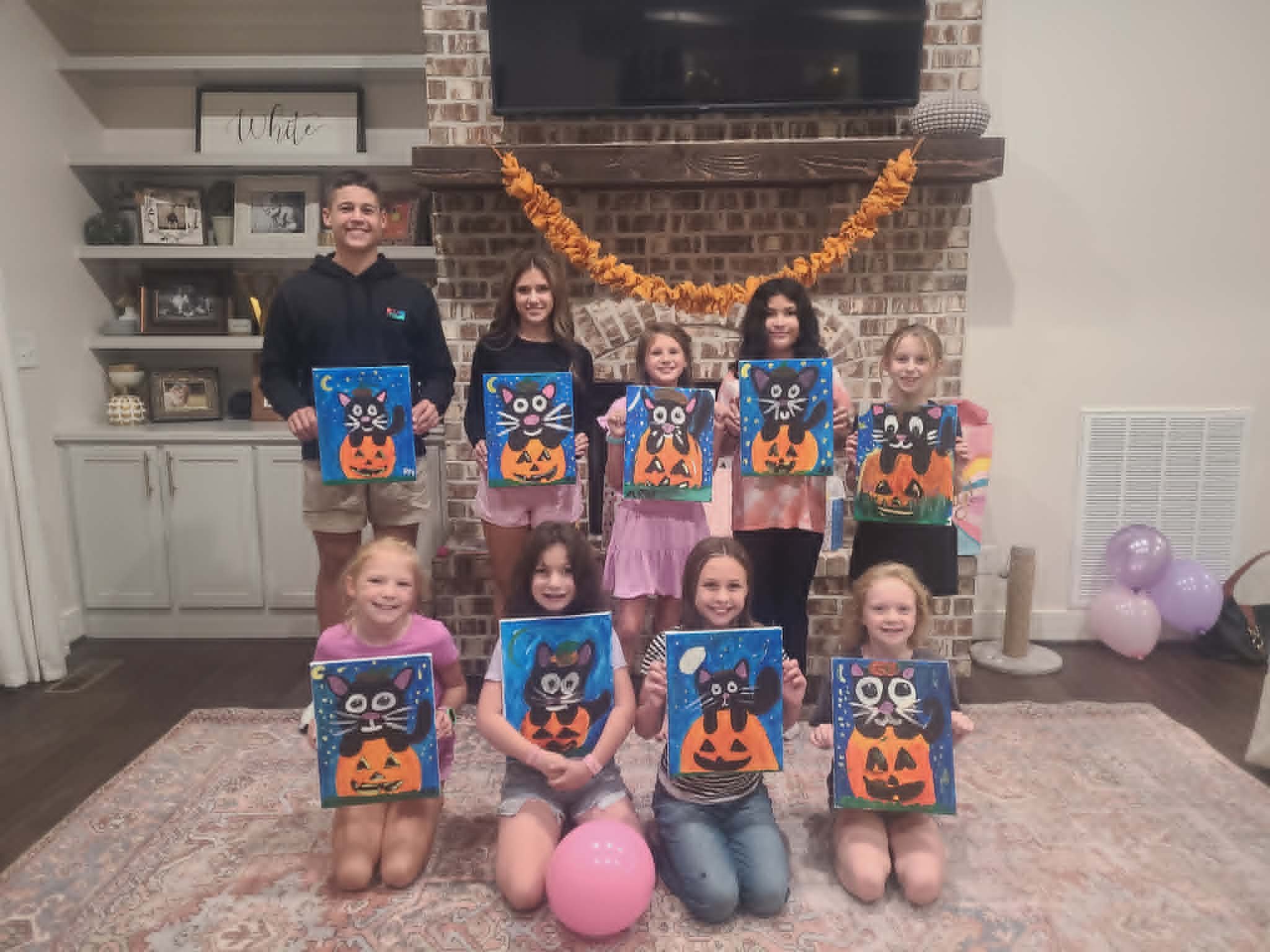 Kids' Halloween paint party group holding up their finished black cat sitting on a jack-o'-lantern paintings with starry blue skies