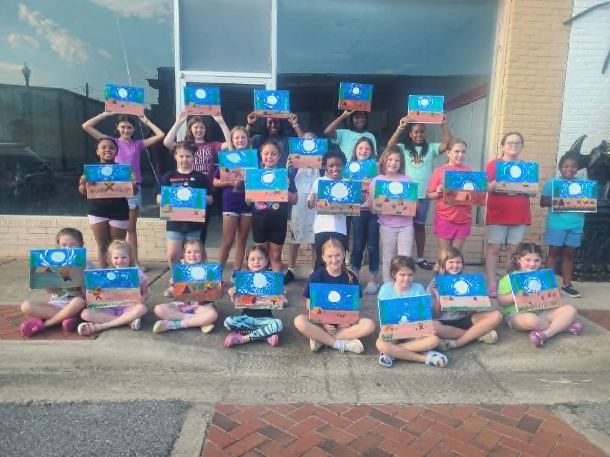 Large group of kids holding up their finished moonlit night beach scene paintings with glowing white moons over sandy shores