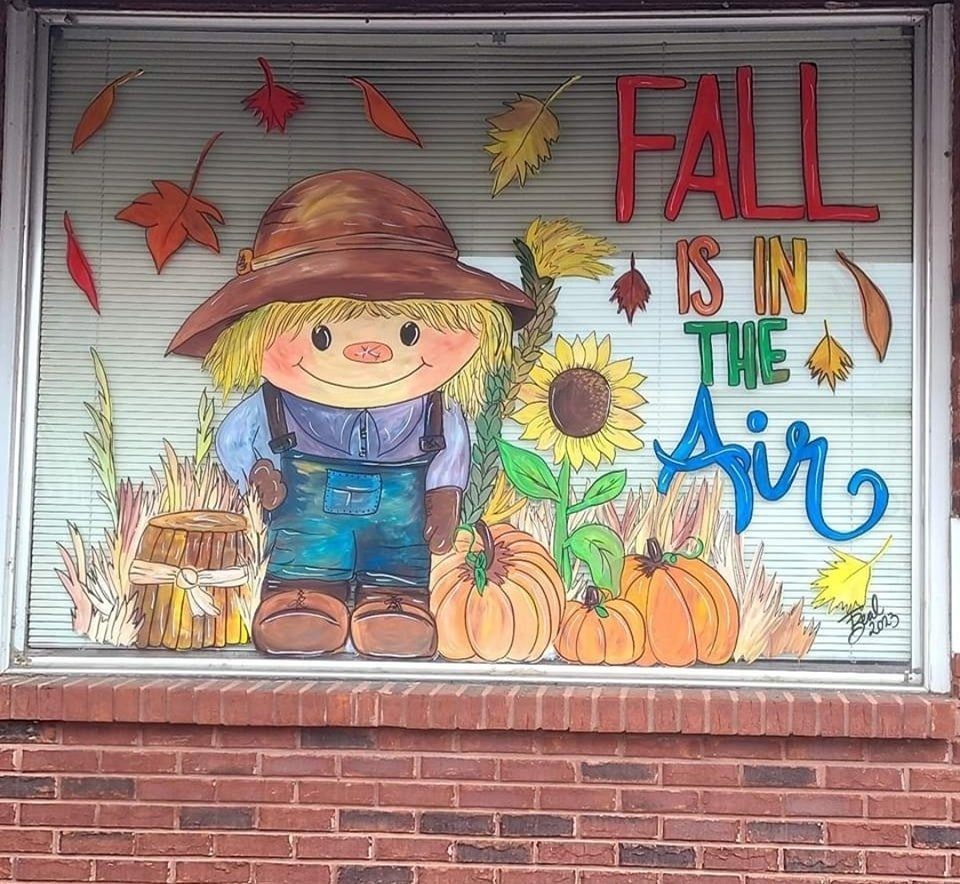 Fall window painting of a friendly scarecrow with sunflowers, pumpkins, and 'Fall is in the Air' lettering