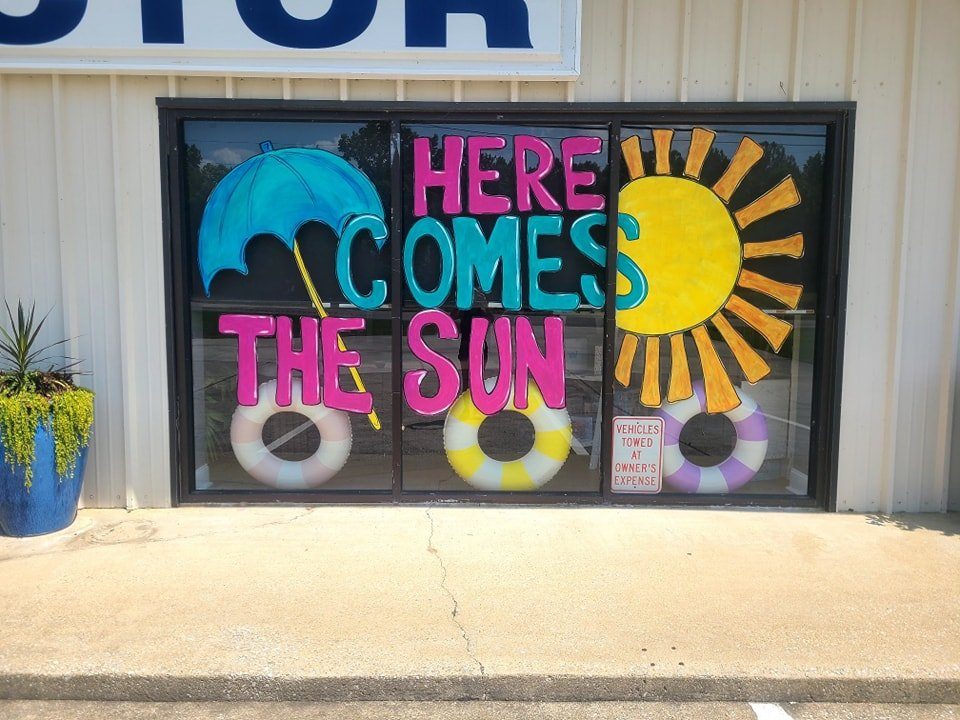 Summer window painting reading 'Here Comes the Sun' with umbrella, sun, and pool floats