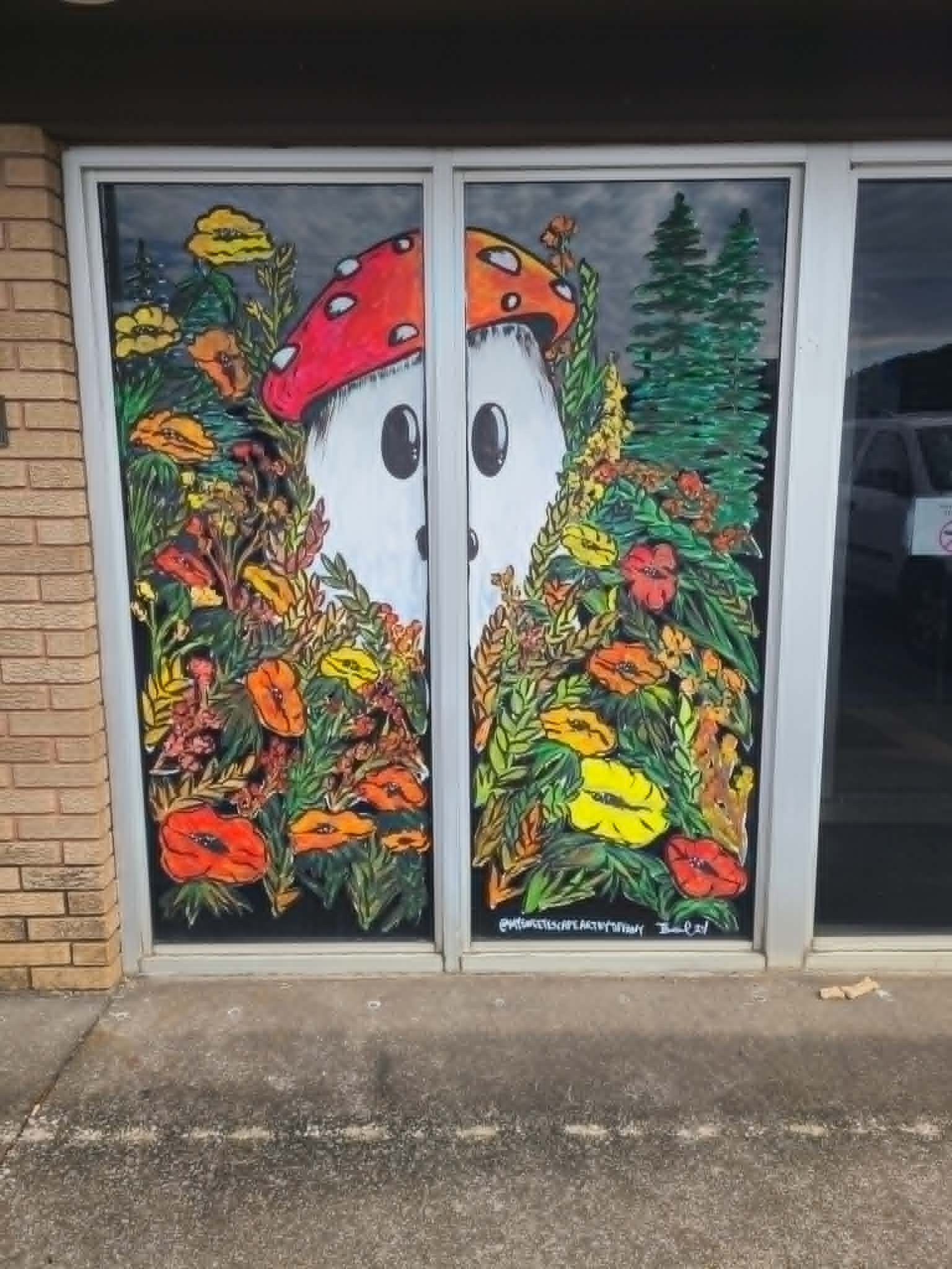 Vibrant cottagecore window painting with twin red-and-white mushrooms peeking out of a colorful wildflower meadow with pine trees