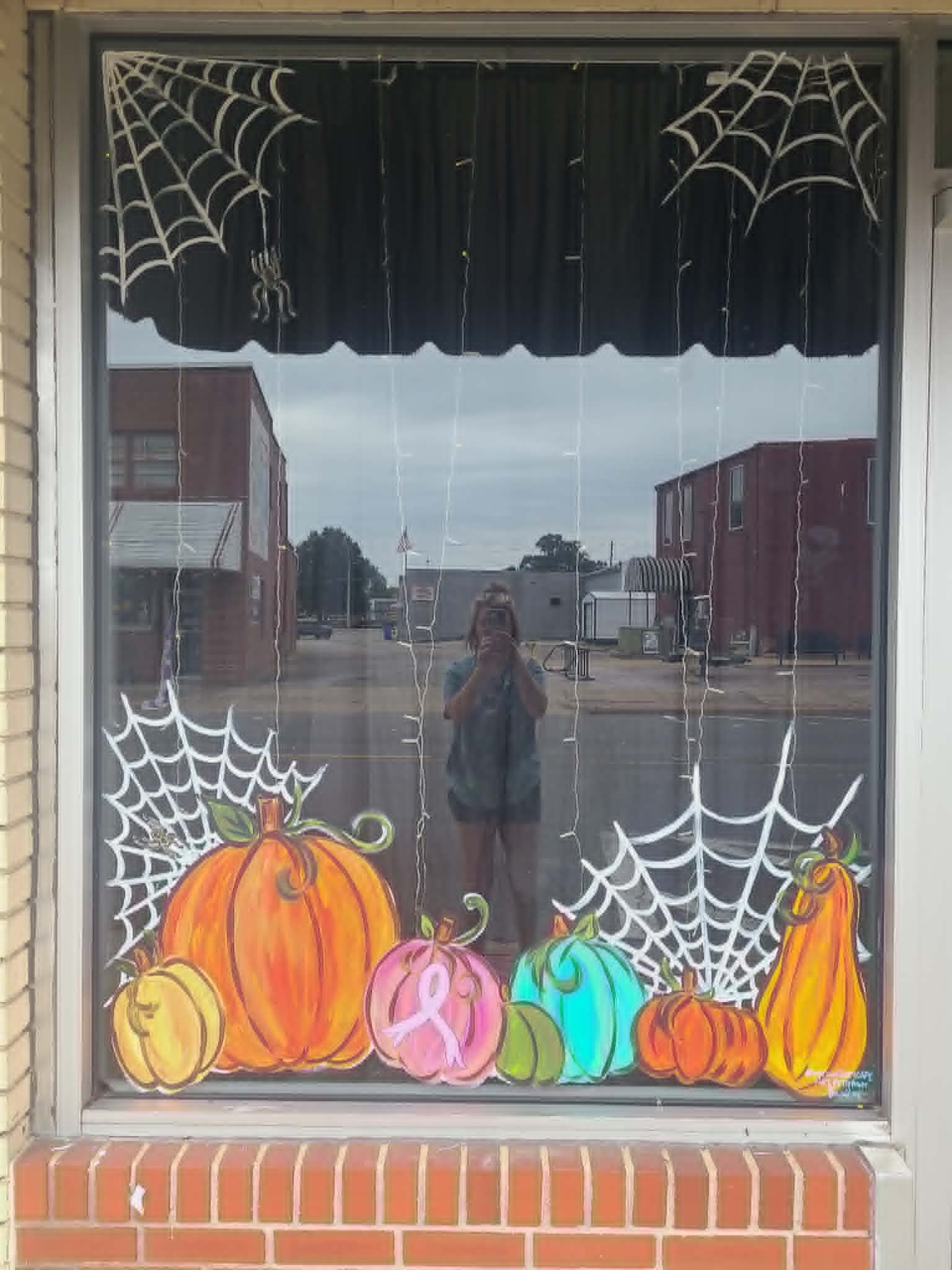Halloween window painting with spider webs, a row of multicolored pumpkins including a pink breast cancer awareness pumpkin, and a small dangling spider