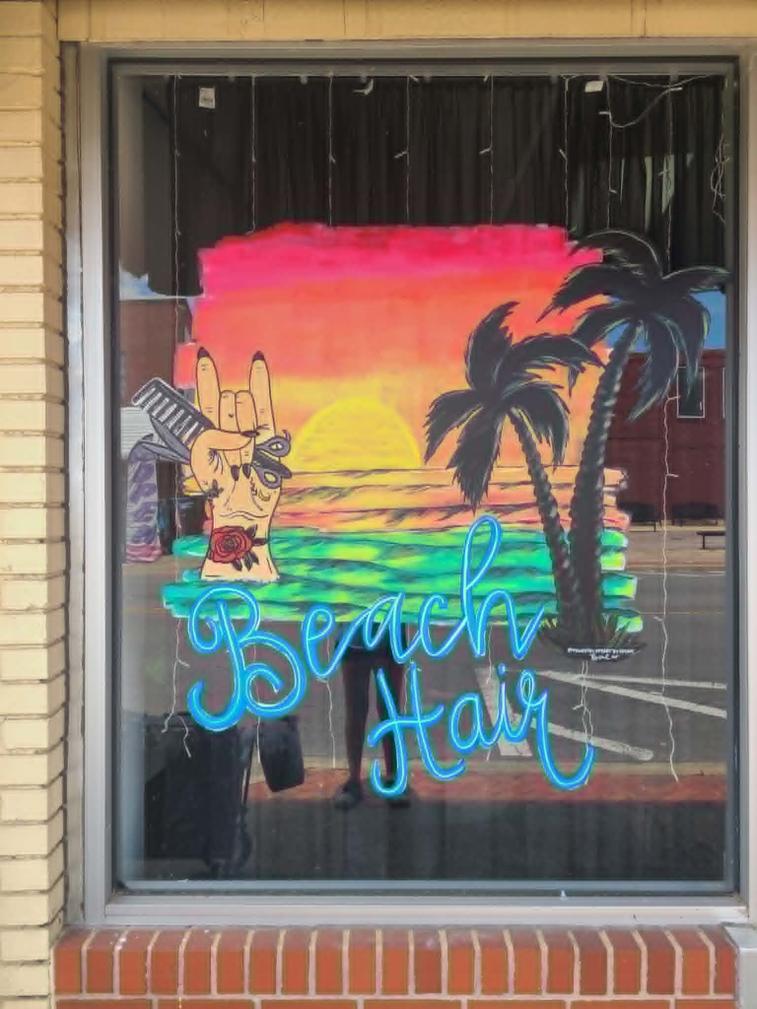 Salon window painting with a tropical sunset, palm trees, a tattooed rock-on hand holding scissors and a comb, and 'Beach Hair' in neon-blue script lettering