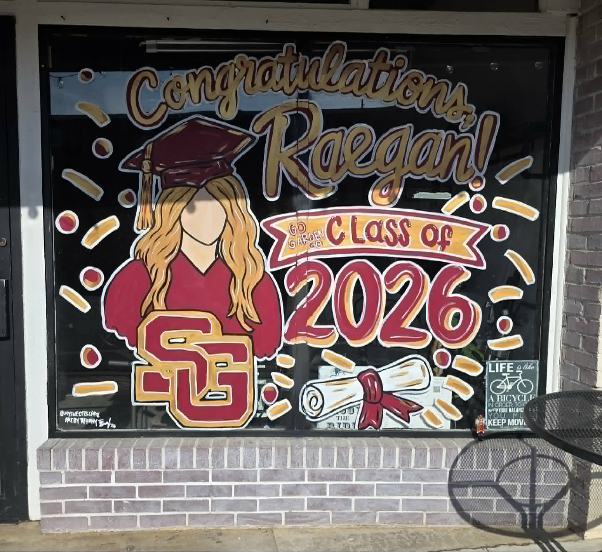 Graduation window painting reading 'Congratulations, Raegan! Class of 2026' with a maroon-and-gold cap, a blonde graduate, the Spring Garden 'SG' logo, and a diploma