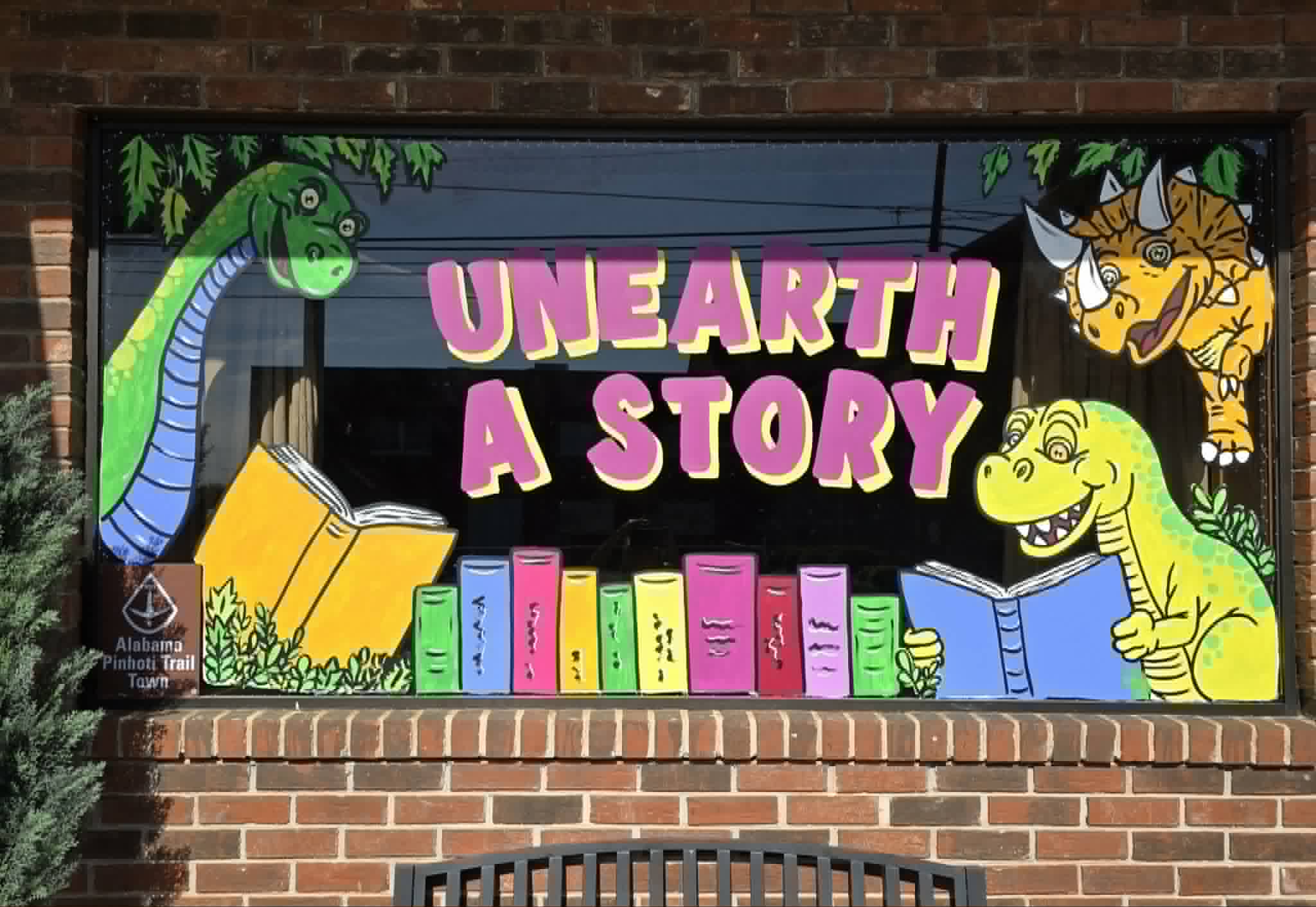 Library window painting reading 'Unearth a Story' with colorful cartoon dinosaurs reading books above a row of bright book spines