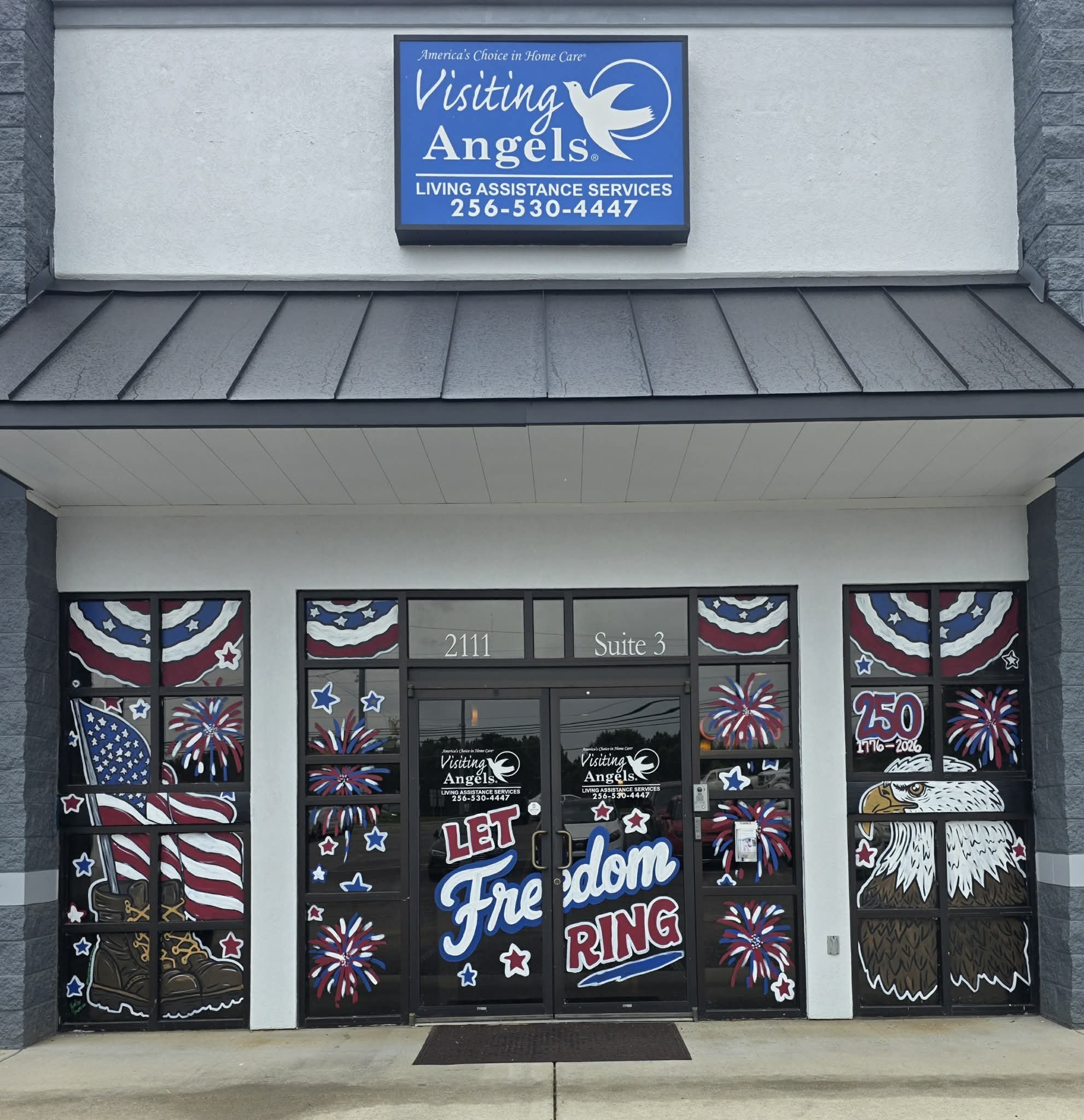 Visiting Angels storefront windows painted with a patriotic 250th anniversary theme — an American flag with combat boots, a bald eagle, fireworks, and 'Let Freedom Ring' lettering