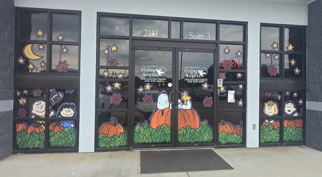 Visiting Angels storefront windows painted with a Peanuts Halloween pumpkin patch — Charlie Brown, Lucy, Linus, Sally, Snoopy in a ghost sheet and Woodstock among orange pumpkins, autumn leaves, stars and a crescent moon