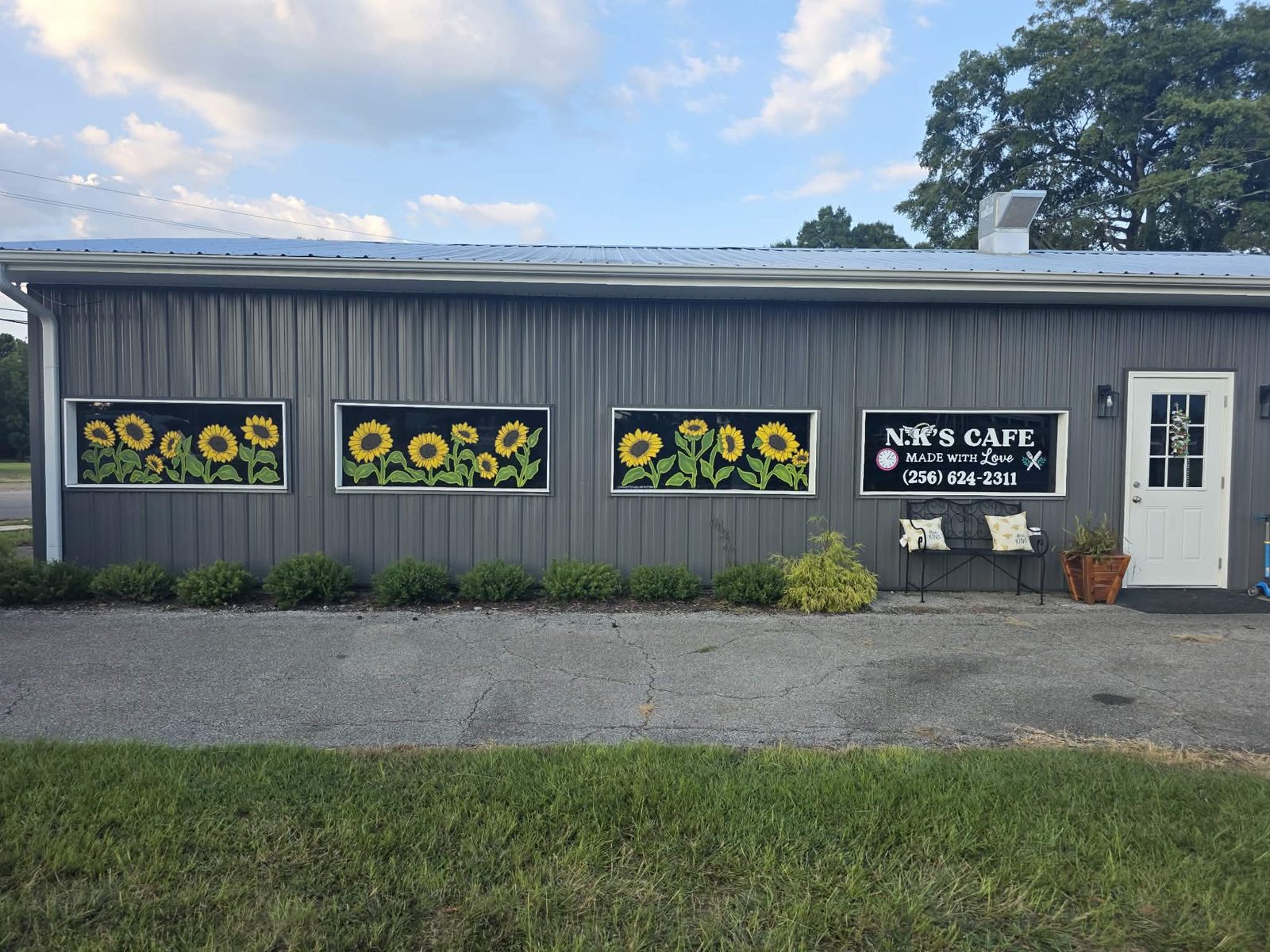 N.K.&rsquo;s Cafe storefront with three long windows painted with rows of bright yellow sunflowers on a black background, beside a painted cafe sign with the phone number