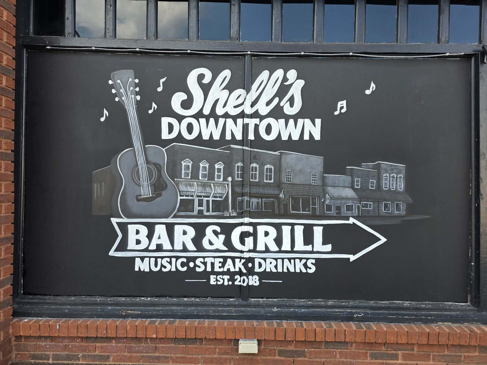 Shell&rsquo;s Downtown Bar & Grill window painted in white on black with an acoustic guitar, a downtown Piedmont streetscape, music notes and a Music, Steak, Drinks arrow sign