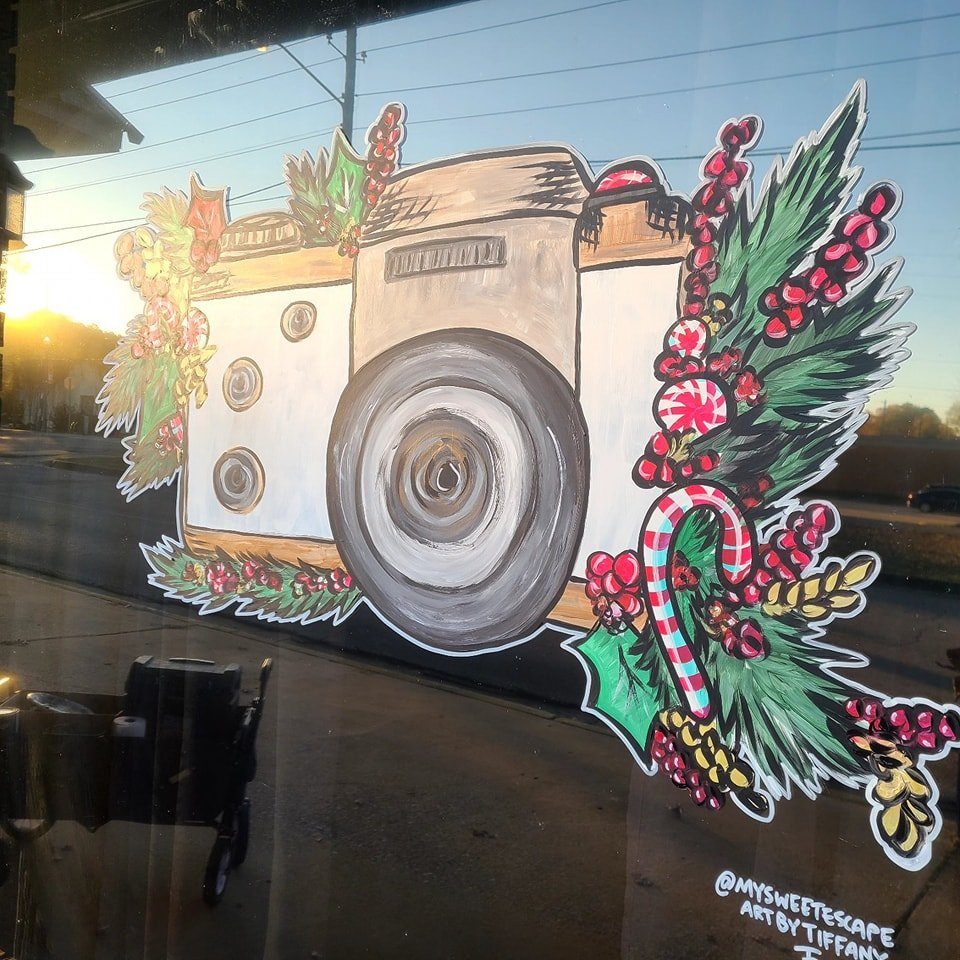 Christmas window painting of a vintage camera surrounded by pine, holly, and candy canes